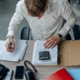 Antreprenor care analizează facturi și un tabel de încasări cu ajutorul unui calculator, la birou