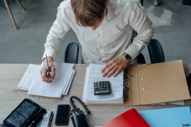 Antreprenor care analizează facturi și un tabel de încasări cu ajutorul unui calculator, la birou